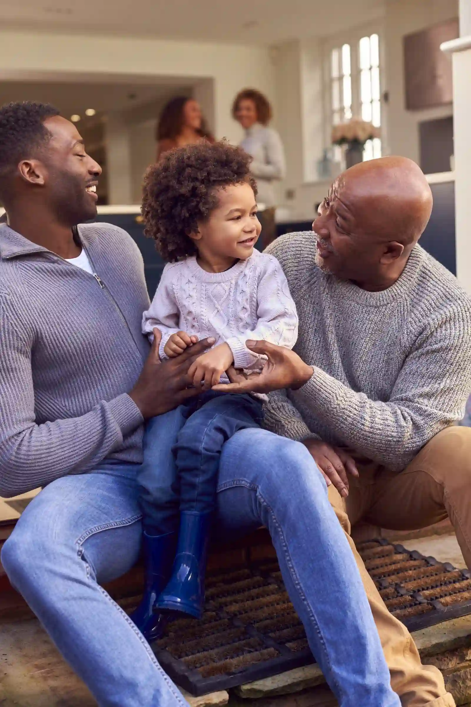 A smiling child sits between two adults indoors, with two people in the background talking.